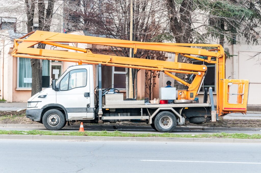 A white truck with an orange boom lift parked on a street, with a worker preparing for maintenance near a building.