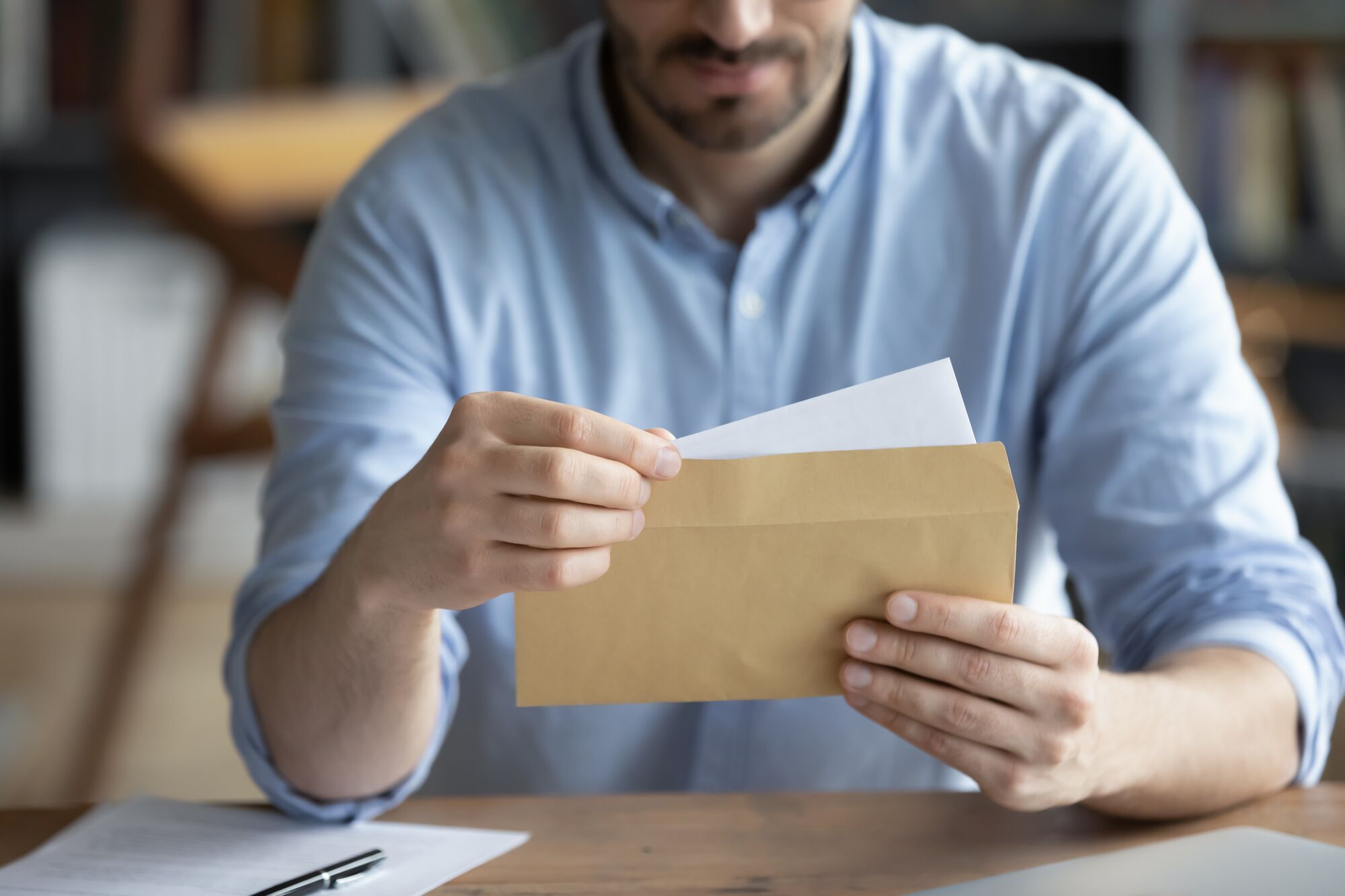 A man in a light blue shirt holds a brown envelope, preparing to open it. Papers are visible, suggesting anticipation or importance of the contents.