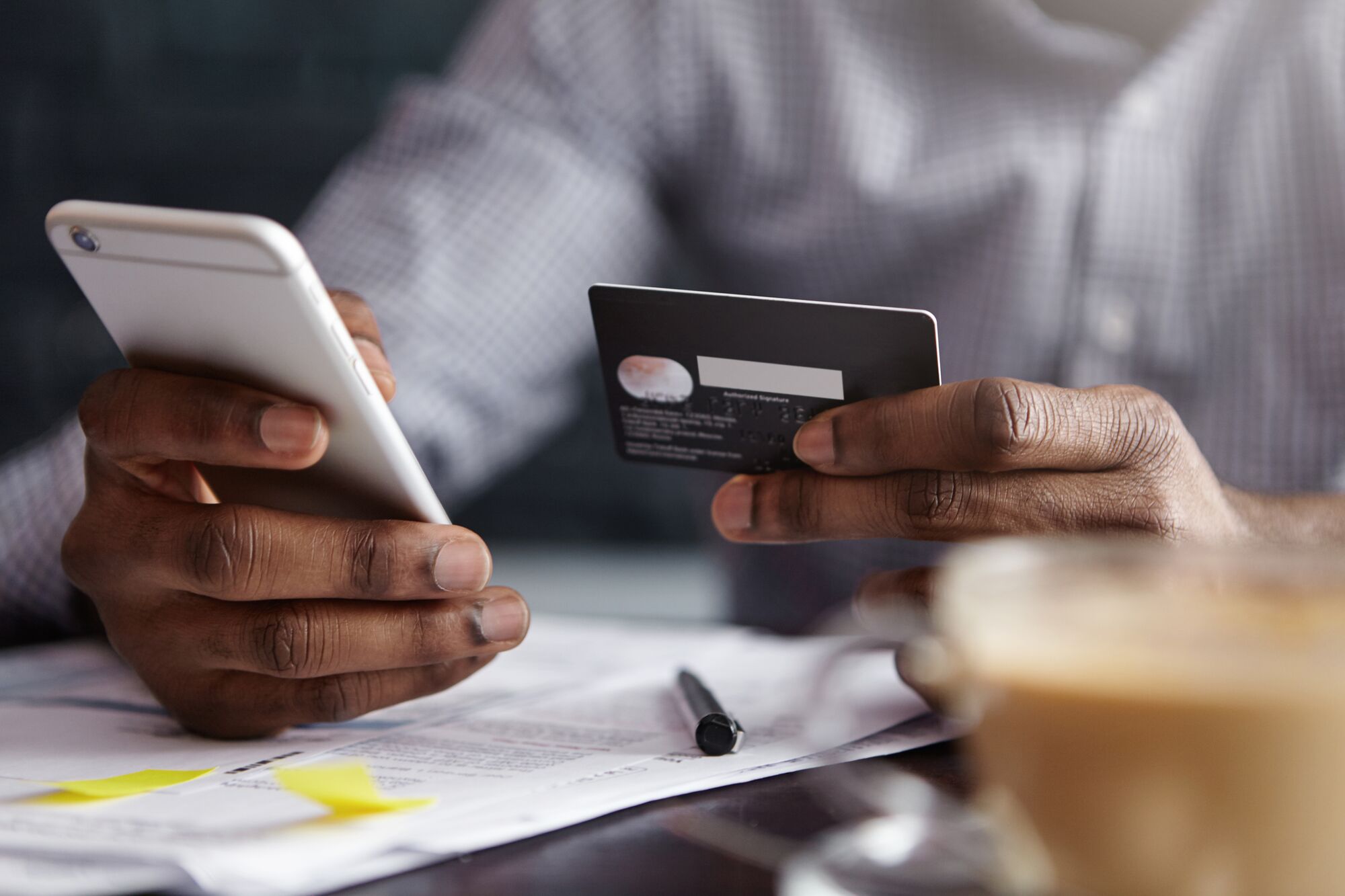 A person holds a smartphone and a credit card while reviewing documents on a table, suggesting an online transaction or payment process.