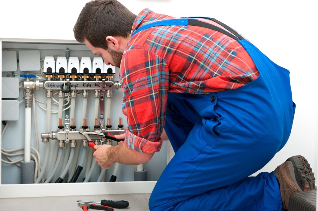 A technician in a plaid shirt kneels beside a plumbing manifold, using pliers to adjust the system, showcasing plumbing maintenance.