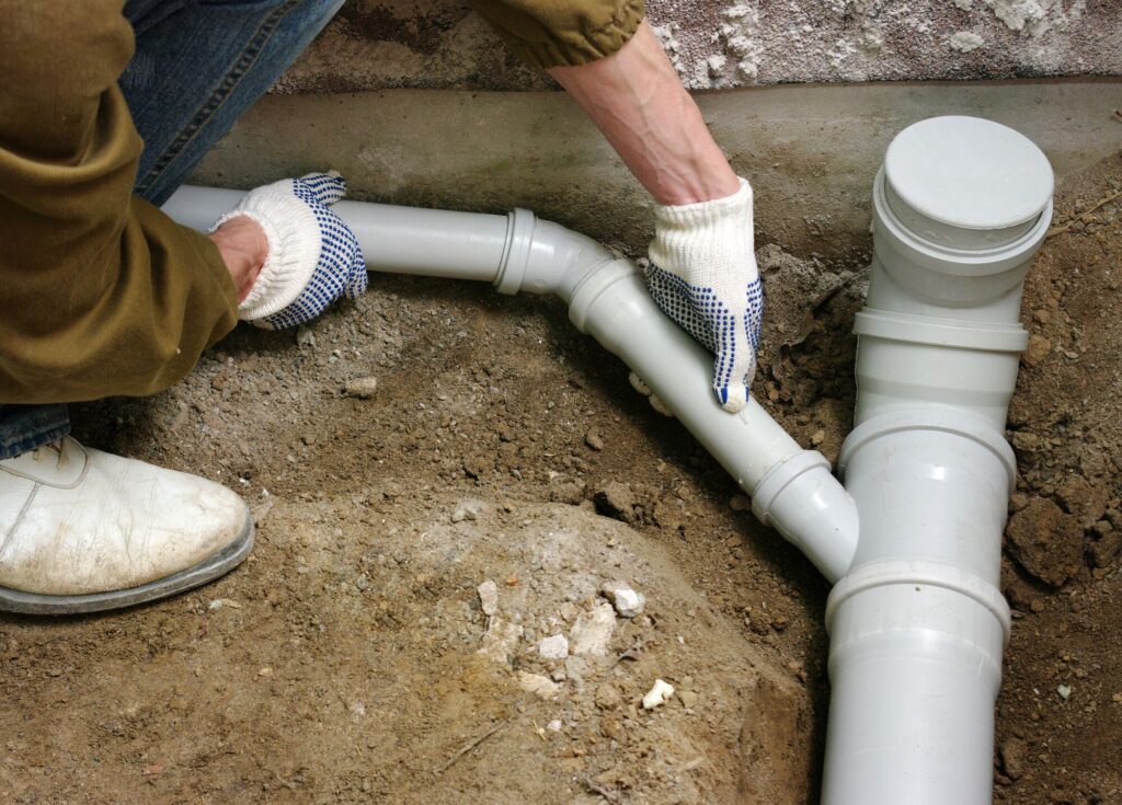 A person in gloves connects PVC pipes in a trench. The image illustrates plumbing work, emphasizing the assembly and installation process.