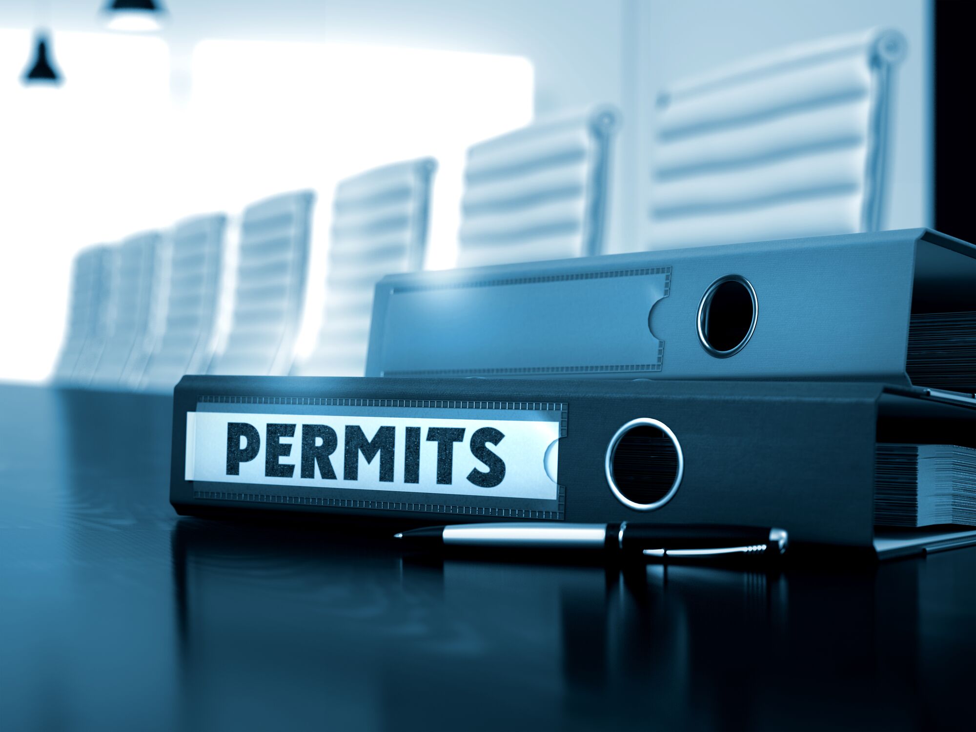 Black binders labeled "PERMITS" are stacked on a conference table, with a pen nearby and a blurred boardroom in the background.