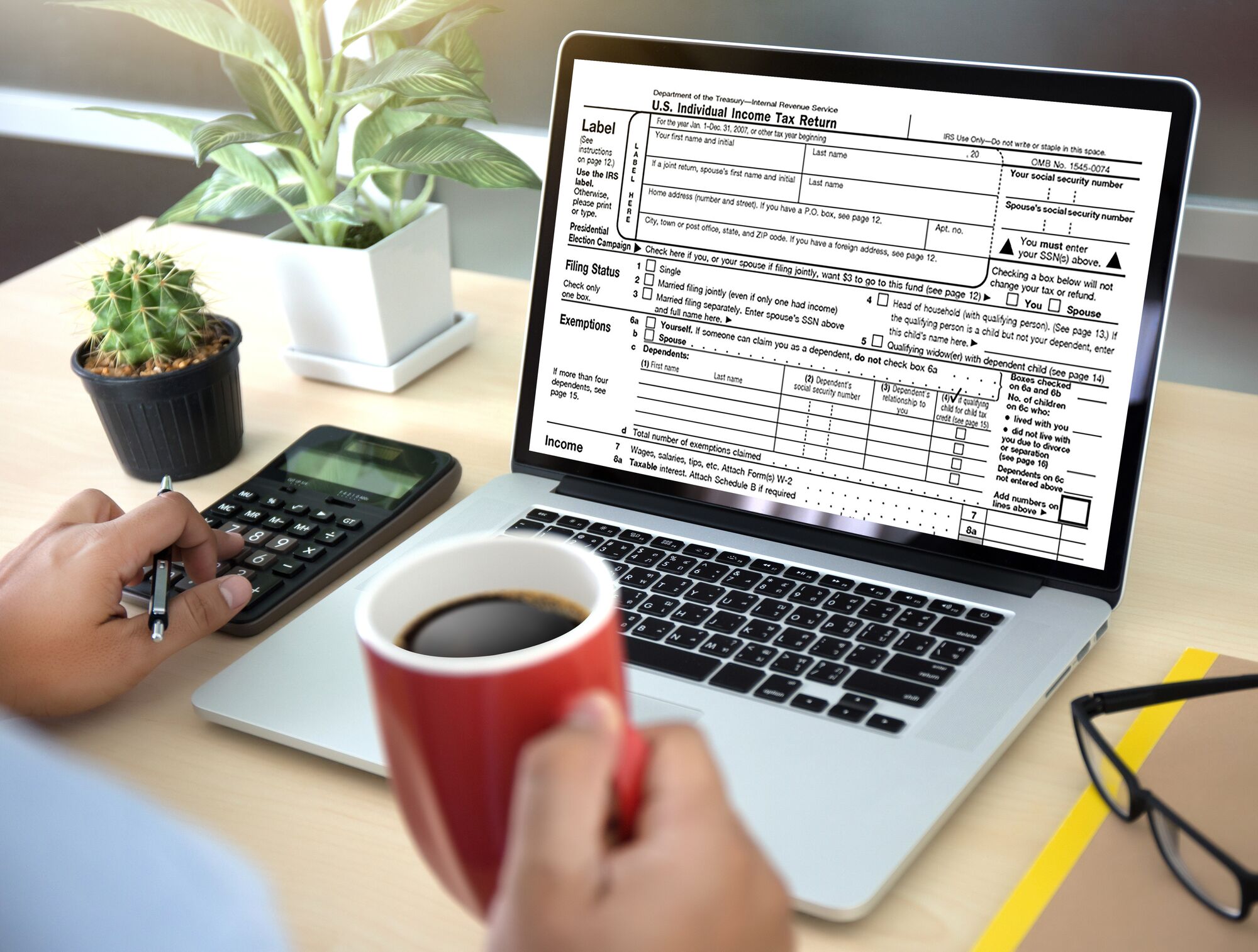 A person holds a coffee cup while using a calculator and filling out a U.S. Individual Income Tax Return on a laptop, surrounded by a plant.