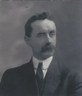 Black and white portrait of a man with a mustache, wearing a suit and tie, showcasing a serious expression and historical attire, relevant for historical context.
