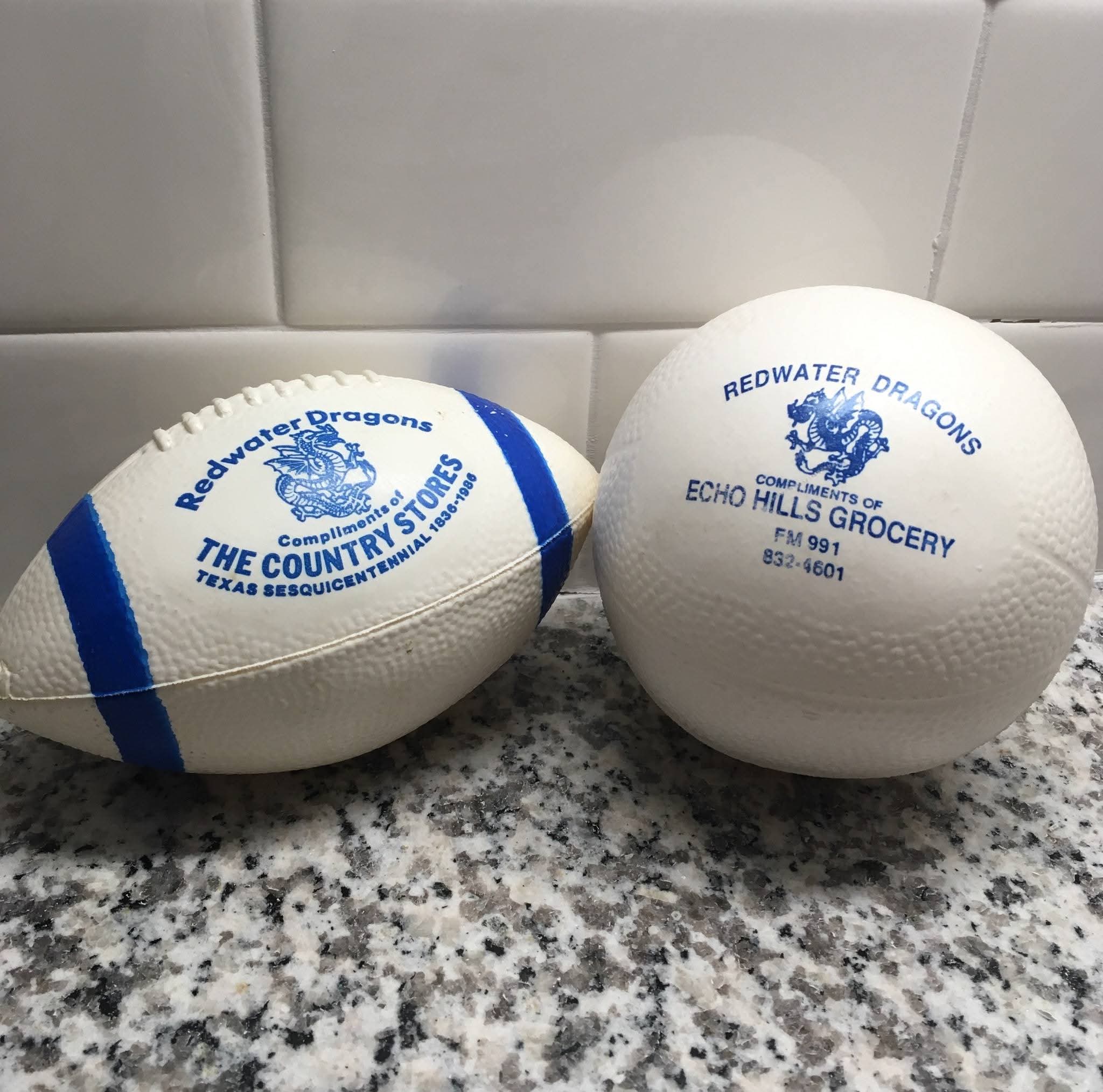 Two promotional balls, one resembling a football and the other a soccer ball, both featuring "Redwater Dragons" and local business names, on a granite surface.