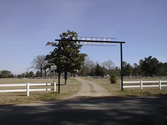 Entrance to Redwater Cemetery features a sign overhead, framed by trees, with gravestones visible in the distance. This image establishes the cemetery's peaceful setting.