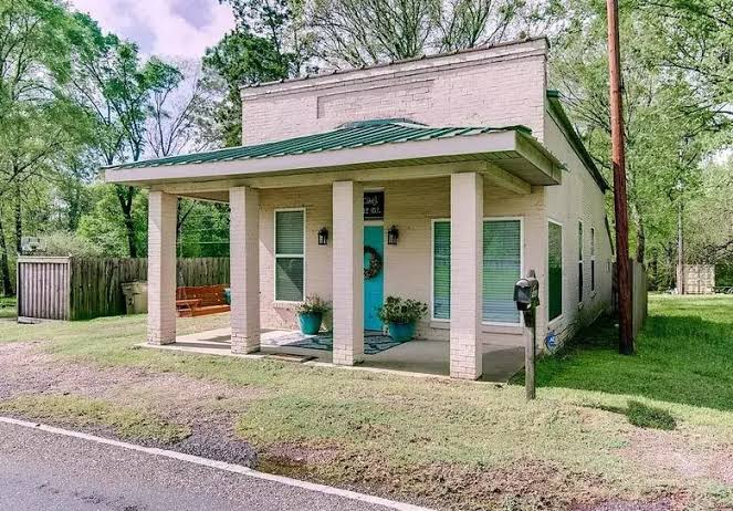 Two-story white building with a green metal roof, featuring a front porch, blue door, and potted plants, surrounded by greenery.