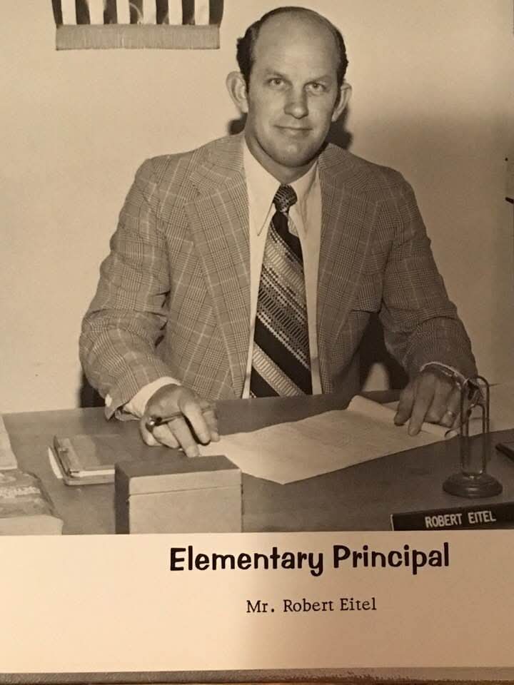 Black-and-white portrait of Mr. Robert Eitel, an elementary principal, seated at his desk with a thoughtful expression. Classroom background includes an American flag.