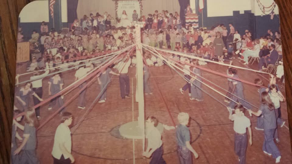 Children participate in a Maypole dance at a school event, surrounded by an audience. Colorful ribbons extend from the central pole, creating a festive atmosphere.