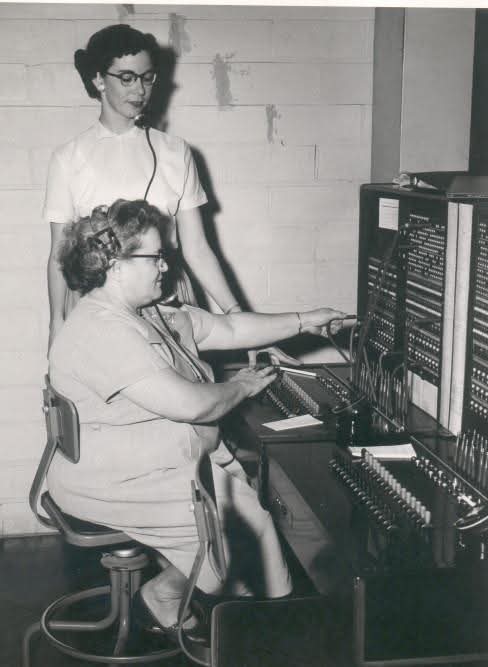 Two women work at an old-fashioned telephone switchboard. One woman, seated, manages the connections, while the other stands behind, observing.